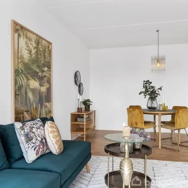 Sofa med pyntepuder står i en lys stue. Rundt sofabord foran, spisebord med fire stole i baggrunden. Dekorativt maleri hænger på væggen. Plante og stearinlys pynter borde.