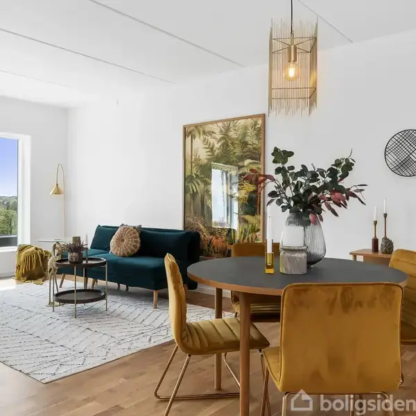 Stue med spisebord og fire stole, blå sofaer og sofabord. En stor plante og vægdekorationer i moderne stil. Vinduer viser udsigt over grønne omgivelser.