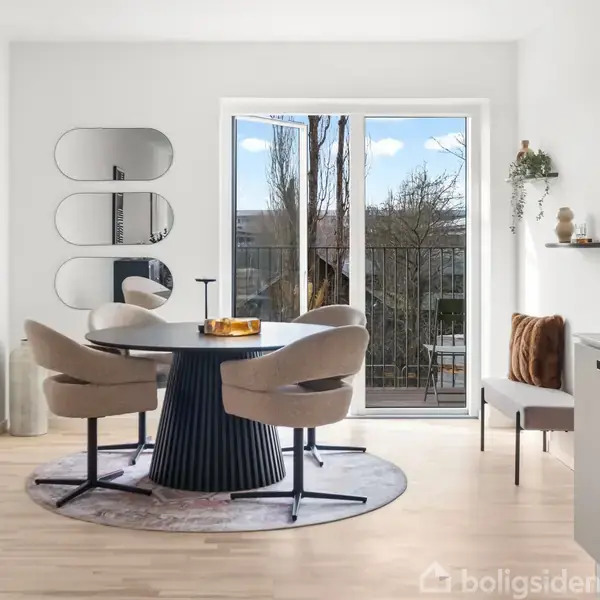 Rundt spisebord med fire stole står i et lyst rum. Et glasdør fører til en balkon, og tre spejle hænger på væggen. Enkel indretning og lyse trægulve.