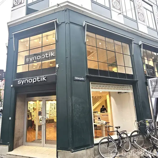 Optikbutik "synoptik" beliggende på et hjørne med store vinduer. Indgangen er åben, og cykler er parkeret udenfor. Skilt på muren viser "Skagade".