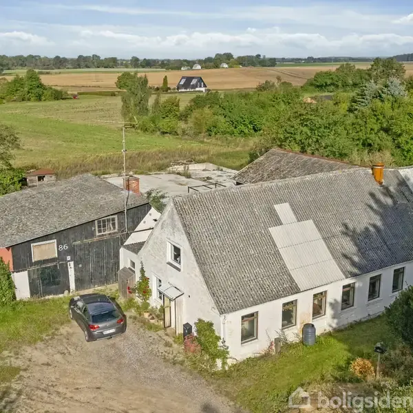 Et gammelt hus med et tilhørende skur står stille i et landligt område omgivet af marker og træer. En bil er parkeret ved siden af. Skuret har nummeret "86" på døren.