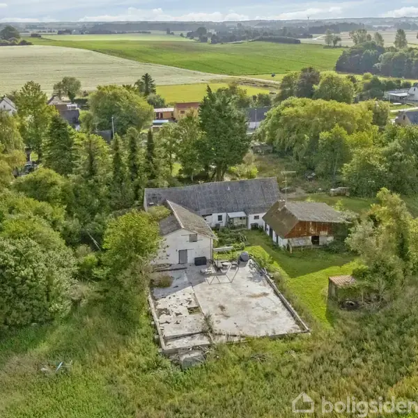 Gårdbygning står tom og forladt, omgivet af grønt landskab og træer. I nærheden ses andre huse og marker i en landlig landsby.