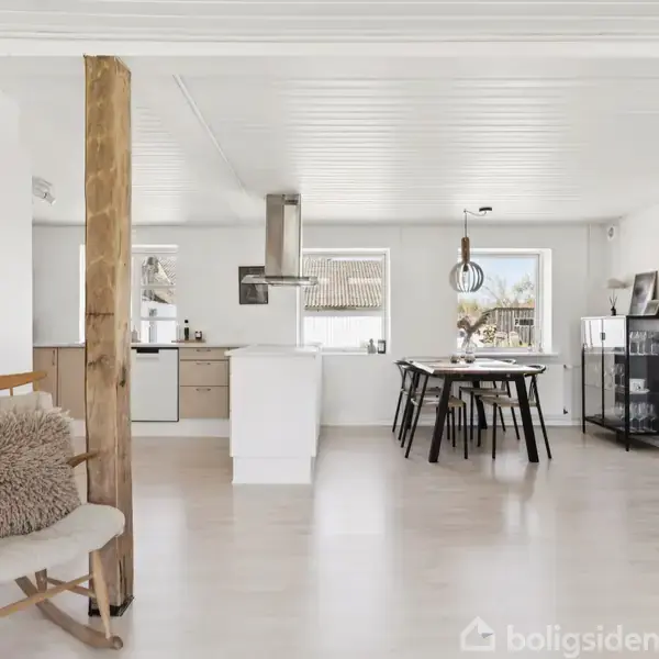 Stue med et træstolpe, lænestol med pude, spisekøkken og brændeovn; lyst og moderne indrettet med hvide vægge og loft, trægulv, spisebord med sorte stole og vinduer med udsigt.