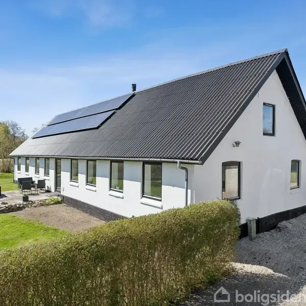 Hvid bygning med sort tag og solpaneler, placeret i en åben have med en stenlagt terrasse i forgrunden. Grusvej og hæk omkranser området under en klar blå himmel.