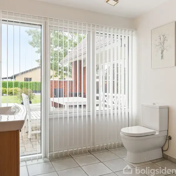 Toilet med lukket låg står ved siden af vinduer med lodrette persienner, med udsigt til terrasse og have. Der er også en håndvask og et brusekabinehjørne i rummet.