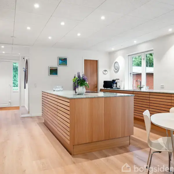 Køkkenø i lysttræ står centralt, omgivet af hvidmalede vægge, kunst på væggene, og spisebord til højre. Store vinduer giver naturligt lys. Trægulv understreger det moderne design.