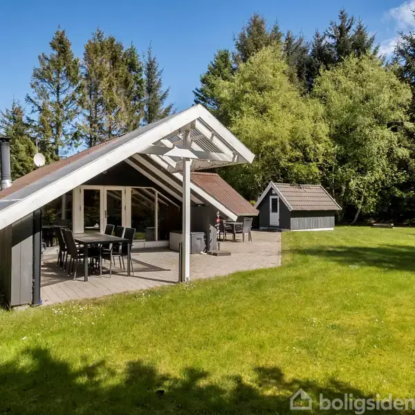 Sommerhus med terrasse og havemøbler står på en velplejet græsplæne omgivet af høje træer og blå himmel. Der er også et lille skur ved siden af hovedbygningen.