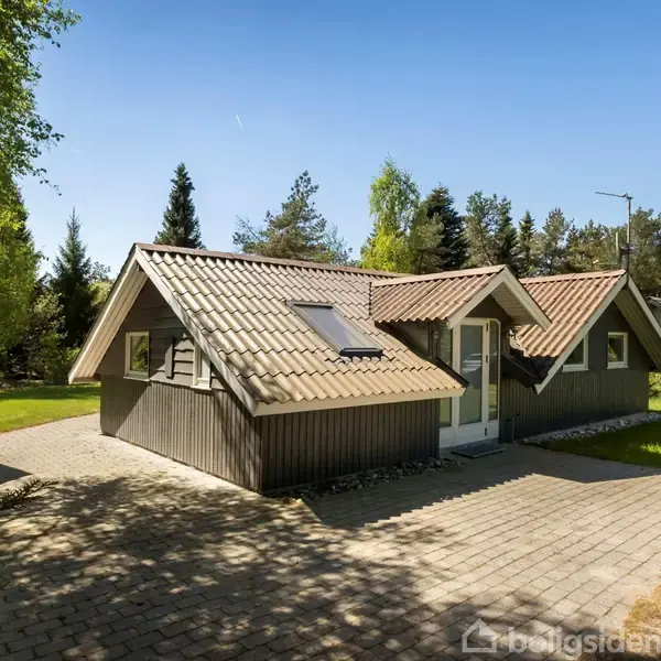 Et sommerhus med træfacade og skråt tegltag står på en flisebelagt indkørsel, omgivet af græs og træer på en solrig dag.