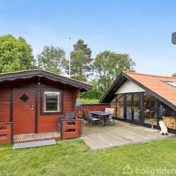 En rød træhytte og en glasvægget bygning står i en grøn have. Et træterrasseområde forbinder de to strukturer. Skyfri blå himmel. Tekst: "Visualisering".
