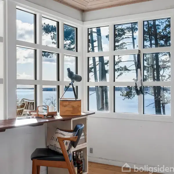 Seng med hvide puder og tæpper står i et lyst rum. Ved siden af er et lille sengebord, en stol med pude, og en fladskærm på et lavt bord. Vindue med udsigt.