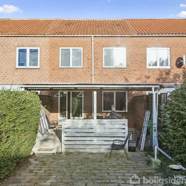 Rækkehus i mursten med rødt tegltag. I forgrunden en flisebelagt terrasse omgivet af buske. Stol og tørrestativ står på terrassen under halvtag, der er fastgjort til huset.