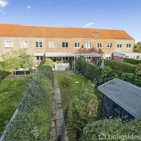 Rækkehus med rødt tegltag står bag en velplejet have. Haven har en gangsti, omgivet af buske og træer, der fører til et skur. Skyfri blå himmel i baggrunden.