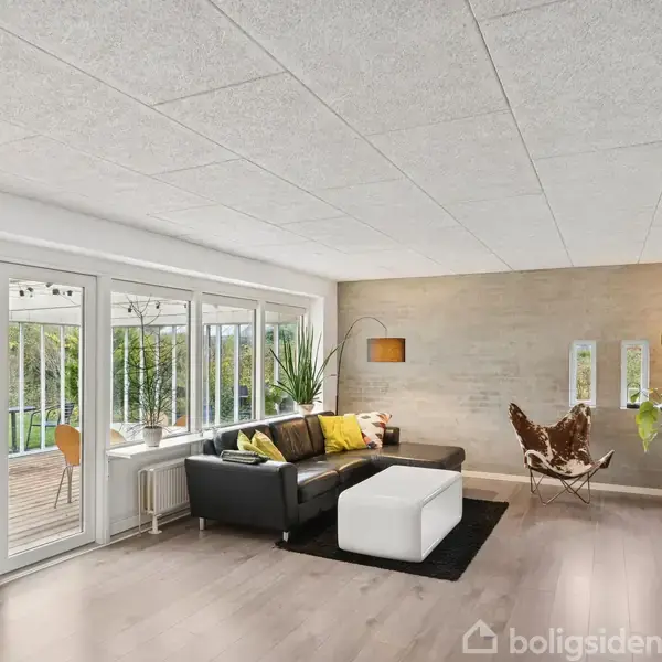 En stue med sort sofa og hvid puf står på et mørkt tæppe. Store vinduer åbner op til en terrasse med udsigt til have.