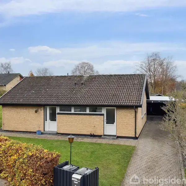 Et enkelthus i gule mursten med mørkt tag står på en grøn græsplæne. Til højre er der en flisebelagt indkørsel, og omgivelserne omfatter hække og træer.