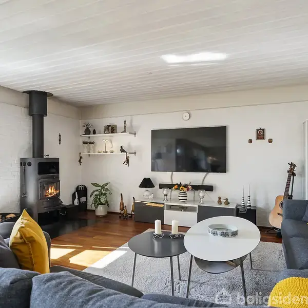 Stue med grå sofa og gule puder, hvor en brændeovn er tændt i hjørnet. Der hænger en fladskærm på væggen, og der står småborde foran sofaen. En guitar står ved siden af et glas skab.