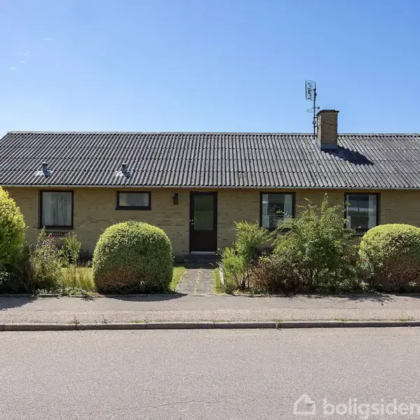 Et gult murstenshus med et gråt tag står stille omgivet af buske og en velplejet forhave på en stille villavej.