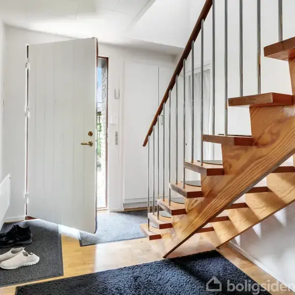 En trætrappe fører opad i en entré med åbentstående hoveddør. På gulvet ligger sko på en måtte. Lys træ og hvidmalede vægge giver et lyst indtryk.