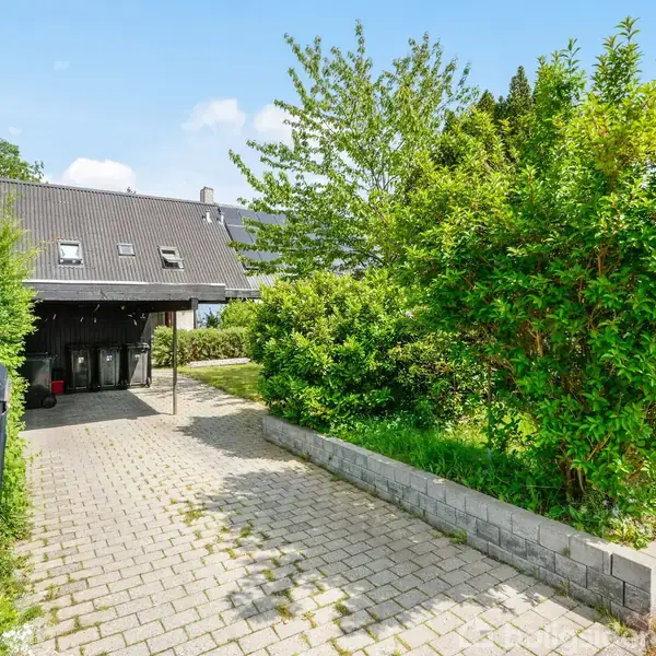 En brolagt indkørsel fører til en carport, omgivet af grønt, tætte buske og et træ til højre. Et sort-tag-hus ses i baggrunden under en blå himmel med få skyer.