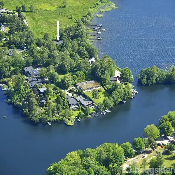 En lille halvø er omgivet af en sø. På halvøen ligger et boligområde med huse blandt træer og haver. Både sejler på søen, og en pil peger på et specifikt hus.