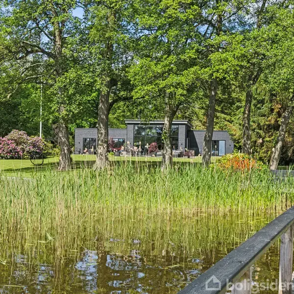 En moderne sort-hus står stille i en skovrig have, omgivet af store træer og blomster. Foran huset er en sø med en træbro i forgrunden.