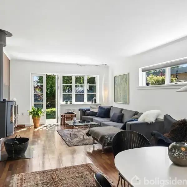 Stue med hjørnesektion placeret foran et stort vindue. En brændeovn står i hjørnet, og der er et rundt spisebord med en lampe hængende over. Rummet er lyst og moderne.