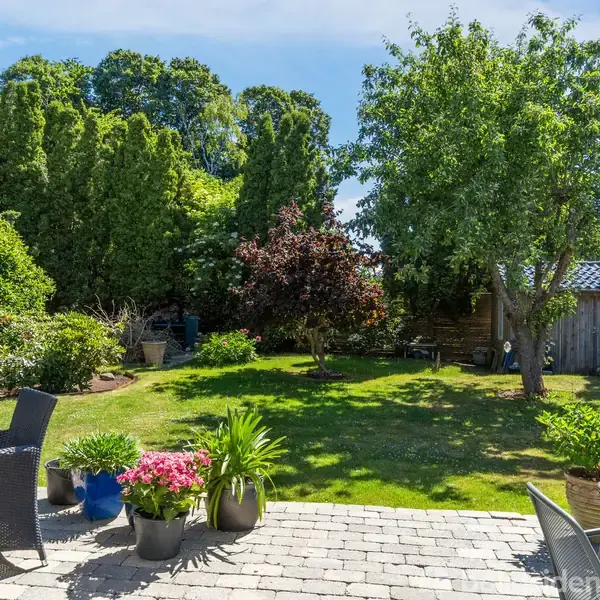 Terrassemøbler står på en flisebelagt terrasse med planter og blomster i potter. I baggrunden ses en grøn have med træer og buske under en klar blå himmel.