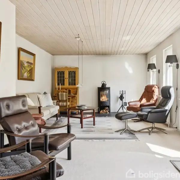 Stue med flere lænestole og en sofa arrangeret rundt et sofabord, pejs i baggrunden. Vægge prydet med malerier, store vinduer til højre med lysindfald, træloft.