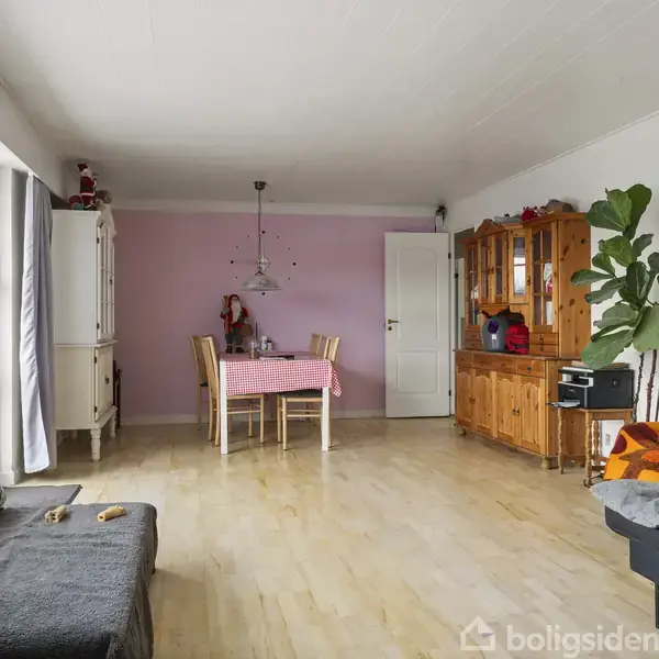 En stue med et bord dækket med en lyserød dug og fire stole. Til højre ses en træskænk og en stor plante. Til venstre er der et stort vindue.