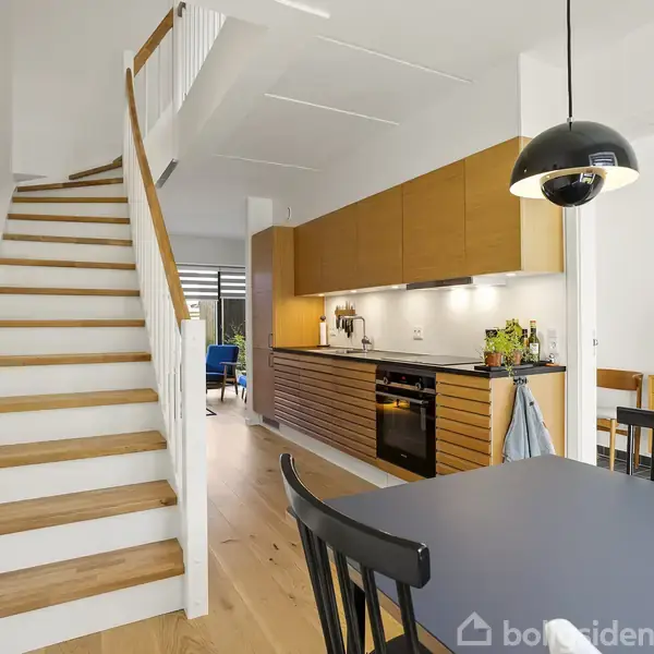 En trappe fører opad i en moderne stueetage med trægulve og et åbent køkken til højre. Køkkenet har træskabe, en sort ovn og hvide vægfliser. En sort pendel hænger over et spisebord.