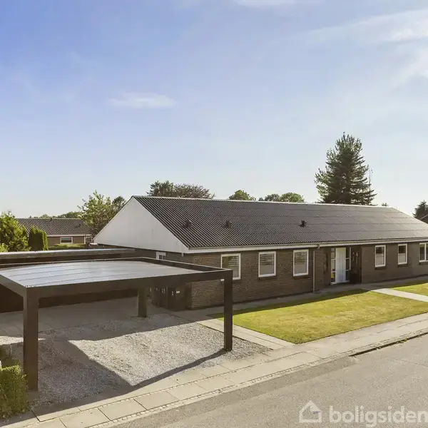 Et enfamiliehus med et sort tag står i et forstadskvarter. Foran huset er en carport og en lille græsplæne. Vejen løber langs ejendommen.