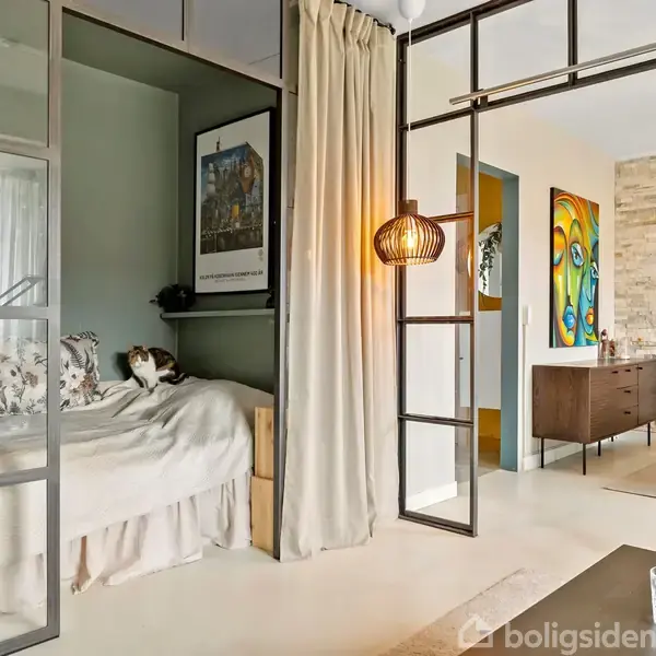 Glaspanel opdeler moderne rum med daybed og pude, hvorpå en kat ligger. I baggrunden ses et spisebord med kunst på væggene og en skænk.