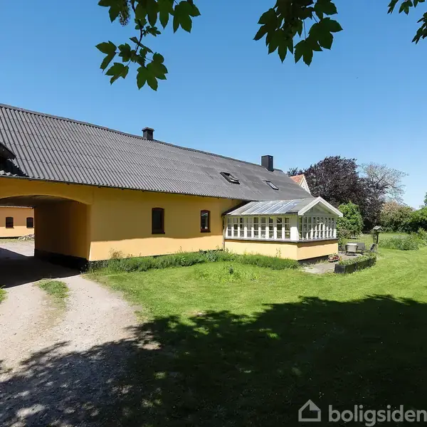 Gult hus med sort tag har en glasveranda, stående i en grøn have med græs og træer omkring. En grusvej fører ind under en port.