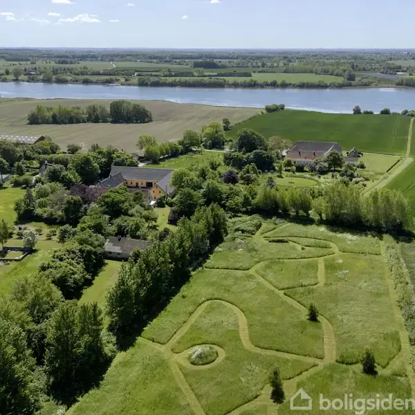 Grøn landskab med snoet vej, omkringliggende træer og marker, flere bygninger i afstand. En stor sø ses i baggrunden under en klar blå himmel.