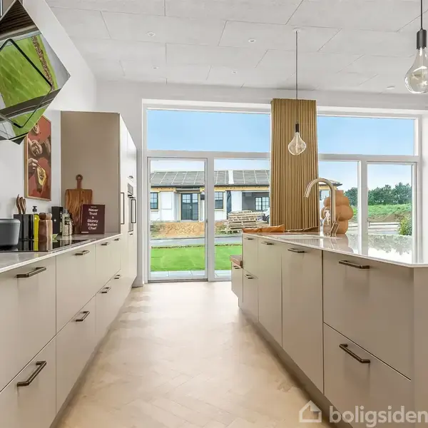 Et moderne køkken med lyse skabe og en køkkenø. Store vinduer i baggrunden giver udsigt til en grøn have, mens diverse køkkenartikler står på bordpladerne.