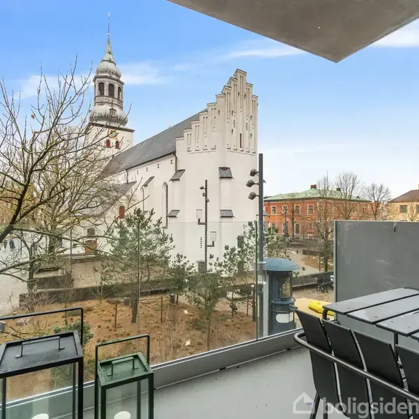 En balkon med et bord og stole, der vender ud mod en kirke med en tårn og givtifuld facade i en bymæssig område med træer og bebyggelse i baggrunden.