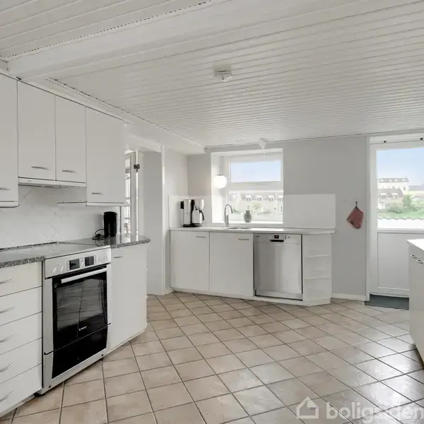Hvidt køkken med moderne apparater, blandt andet ovn og opvaskemaskine, placeret i et rum med beige flisegulv og hvidt loft, omgivet af lyse vægge og vinduer.