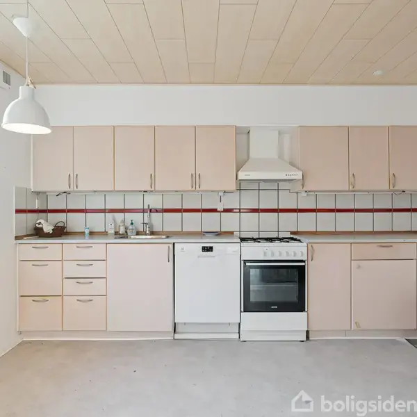 Køkken med beige skabe og skuffer, gaskomfur, ovn og opvaskemaskine under emhætte, arrangeret mod en hvid væg med rød-hvid flisebagvæg, belyst af en loftlampe.