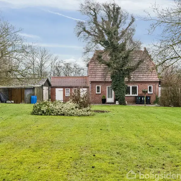 Et rødt murstenshus står stille omgivet af en stor, grøn græsplæne med spredte træer og buske. I baggrunden ses en klar himmel og flere afsides træer.