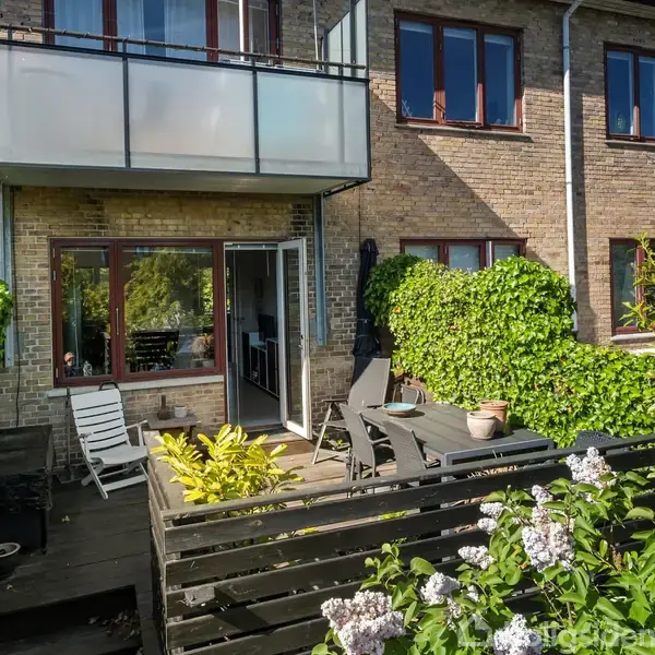 Terrasse med havemøbler, omgivet af grønne planter og buske, foran en murstensbygning med altan og store vinduer. Møblerne står på trædæk, og døren til huset er åben.
