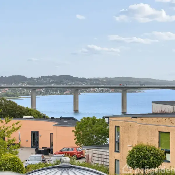 En bro strækker sig over en blå fjord, omgivet af frodige grønne områder og moderne bygninger i et boligområde, med bløde skyer på himlen i baggrunden.