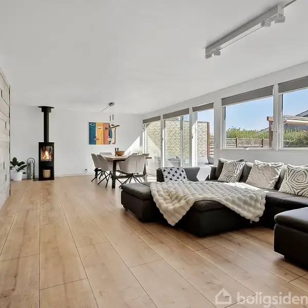 Fladskærms-tv hænger på væggen; sort sofa med tæpper står i stue med trægulv. Spisebord er i baggrunden ved brændeovn. Store vinduer giver udsigt til have.