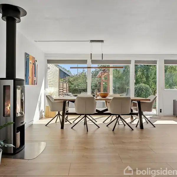 Pejs i et lyst rum opvarmer stille, mens en moderne spisebordgruppe står ved store vinduer, der viser en frodig have. En grøn plante pynter gulvet ved siden af pejsen.