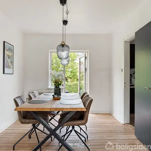 Et spisebord med seks stole og dækkeservietter står i et lysfyldt rum. En lampe hænger over bordet, ved siden af en åben dør og to billeder på væggen.