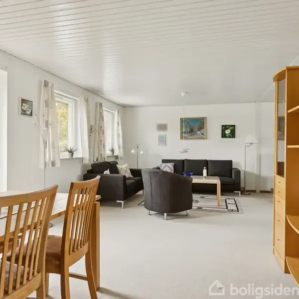 Stue indrettet med træmøbler og sorte sofaer, placeret i et lyst rum med flere vinduer, hængende gardiner, og kunst på væggene. En spisebord og vitrineskab står fremme.