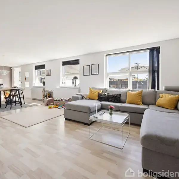 Grå sofa med puder ved glasbord i lys stue. Spisebord i baggrunden, store vinduer lader lys strømme ind. Trægulv og minimalistisk indretning skaber en moderne atmosfære.