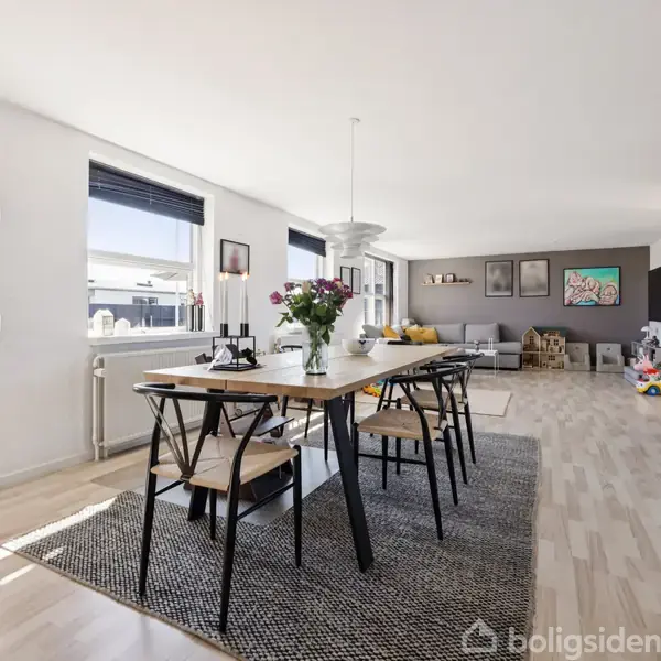Spisebord med blomster i vase står på tæppe. Omgivet af stole og har udsigt til en stue med sofaer, kunstværker på væggen og legetøj i baggrunden.