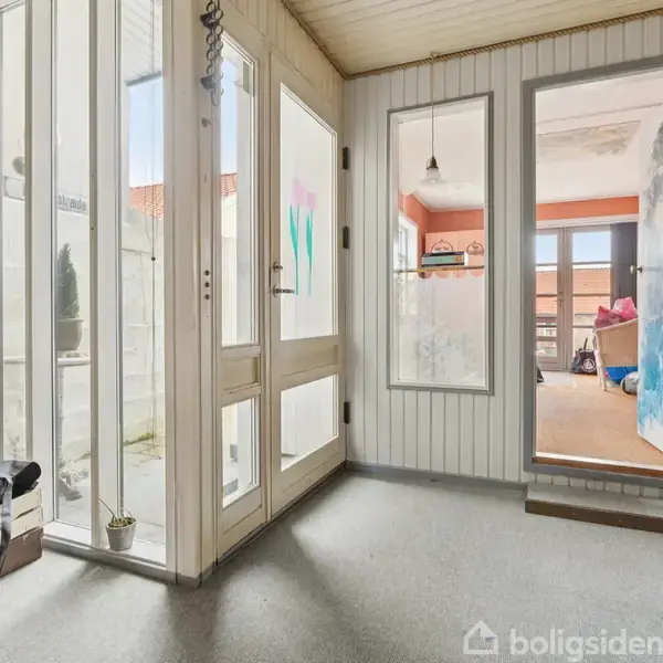 Trappe fører opad, flankeret af gelænder; sofa står under trappe. Billede viser indgang til andet rum, med skrivebord og rød gardin. Kasser og bord til højre.