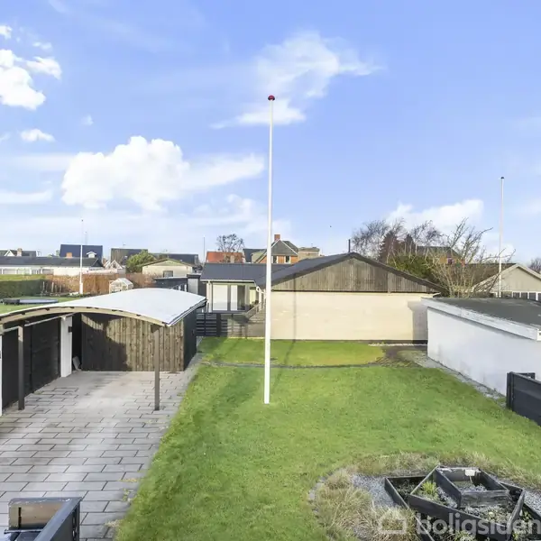 Flagstang står i en grøn have, omkranset af træhegn. Til venstre ses en flisebelagt sti og en carport. Huse ses i baggrunden under en blå himmel med skyer.