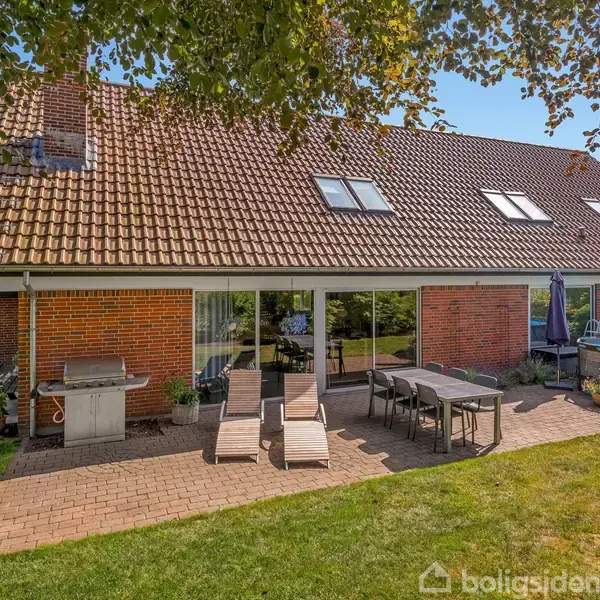 Et rødt murstenshus har en terrasse med liggestole og et spisebord. Der er en grill til venstre. Omkring er der græsplæne og grønne buske under en blå himmel.