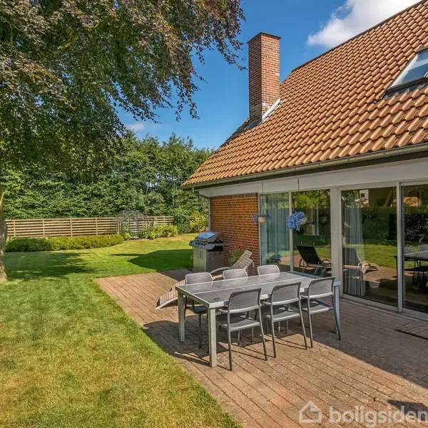 Et bord med seks stole står på en terrasse af fliser ved siden af et moderne hus med rødt tegltag. En stor træ i grøn have omgiver området på en solrig dag.